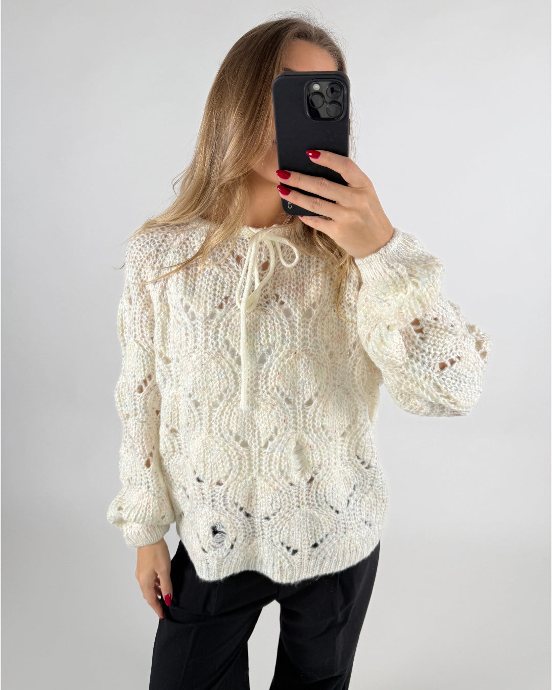 En person tager et spejlbillede iført en hvid kabelstrikket sweater og sorte bukser. Personen holder en sort smartphone, mens baggrunden er neutral og lys.