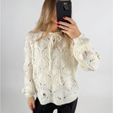 En person tager et spejlbillede iført en hvid kabelstrikket sweater og sorte bukser. Personen holder en sort smartphone, mens baggrunden er neutral og lys.