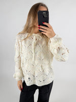 En person tager et spejlbillede iført en hvid kabelstrikket sweater og sorte bukser. Personen holder en sort smartphone, mens baggrunden er neutral og lys.