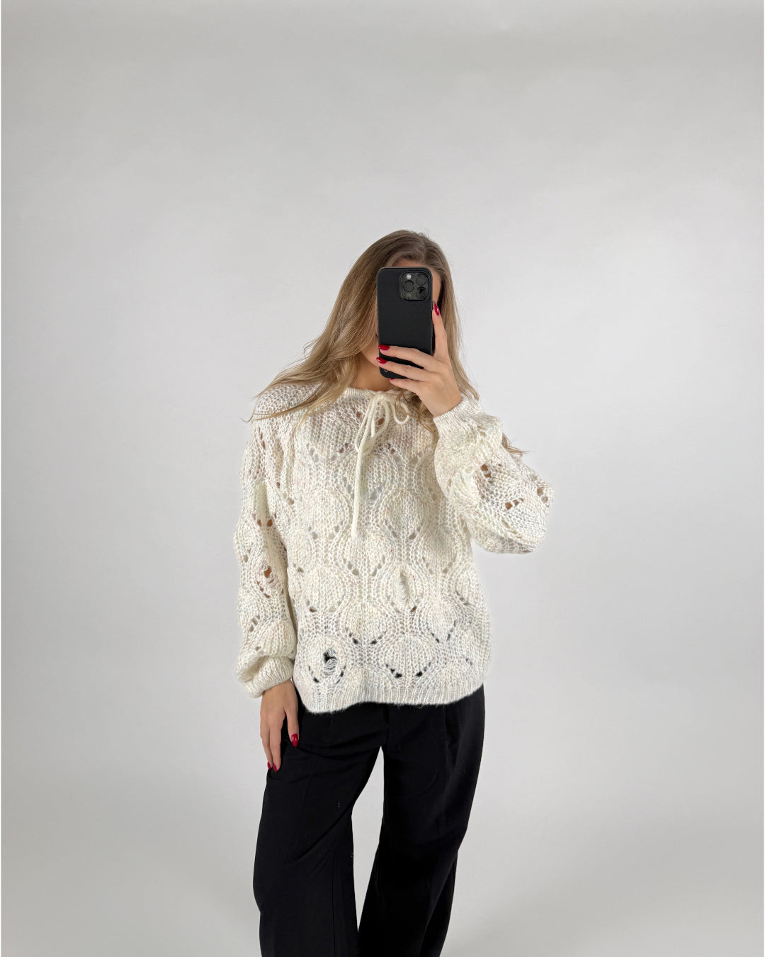 Person tager selfie i spejl iført en hvid strikket sweater med hulmønster og sorte bukser, på en neutral baggrund.