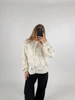 Person tager selfie i spejl iført en hvid strikket sweater med hulmønster og sorte bukser, på en neutral baggrund.