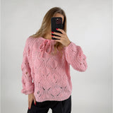 En person iført en lyserød strikket sweater tager en selfie med en mobiltelefon. Personen står i et minimalistisk, lyst rum og har sort tøj på.