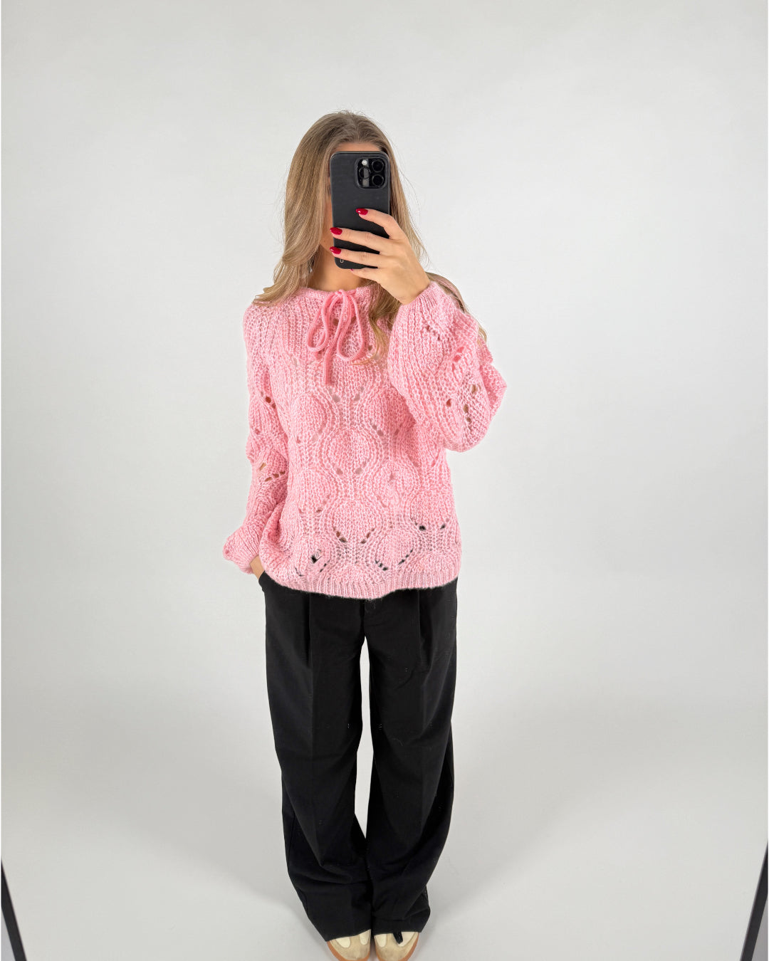 En person iført en lyserød strikket sweater tager et spejlfoto med en smartphone. Personen står i et hvidt rum og bærer sorte bukser og beige sko.