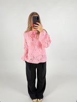 En person iført en lyserød strikket sweater tager et spejlfoto med en smartphone. Personen står i et hvidt rum og bærer sorte bukser og beige sko.