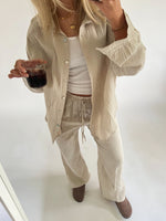 En person iført beige sæt drikker fra et glas. Personen står i et lyst rum med hvid baggrund og brune sko.