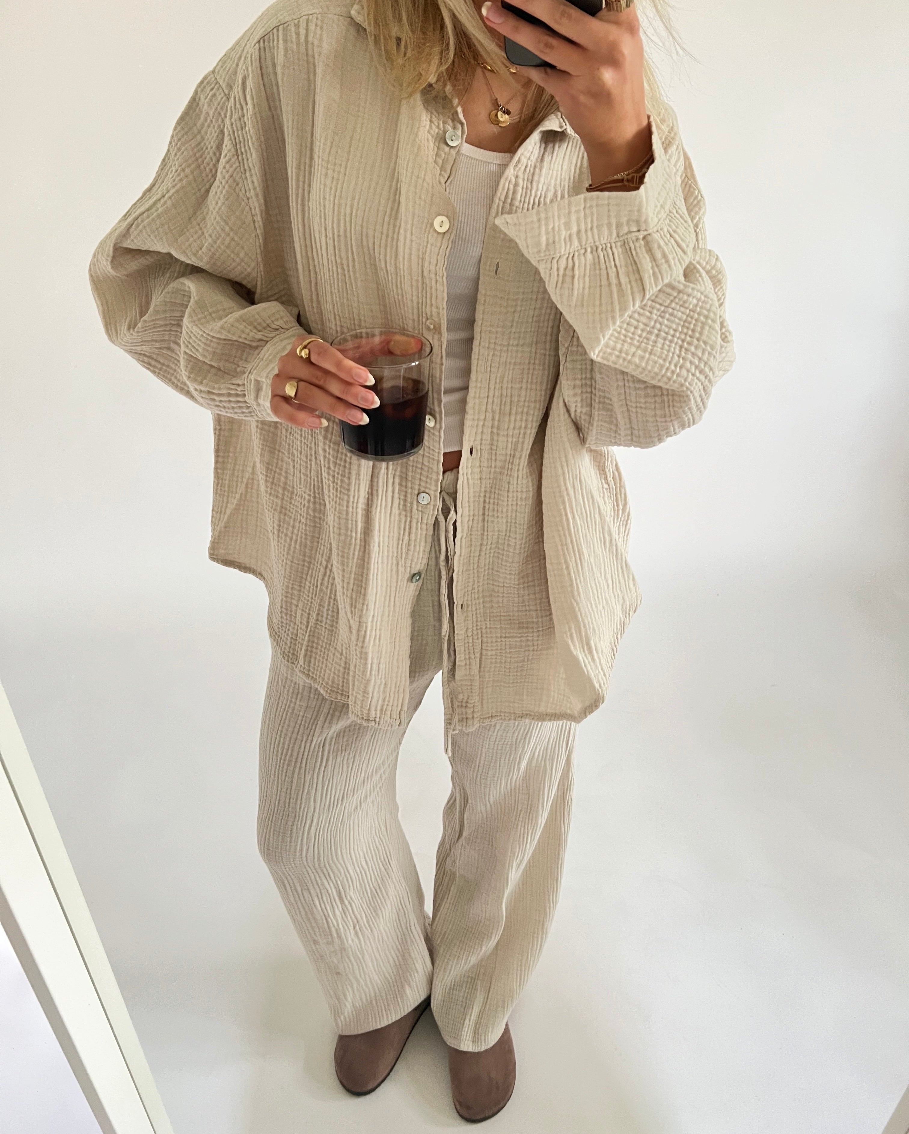 En person iført løstsiddende lyse beige tøj drikker fra et glas. Personen holder en smartphone, står i et enkelt, lyst rum og har støvler på.