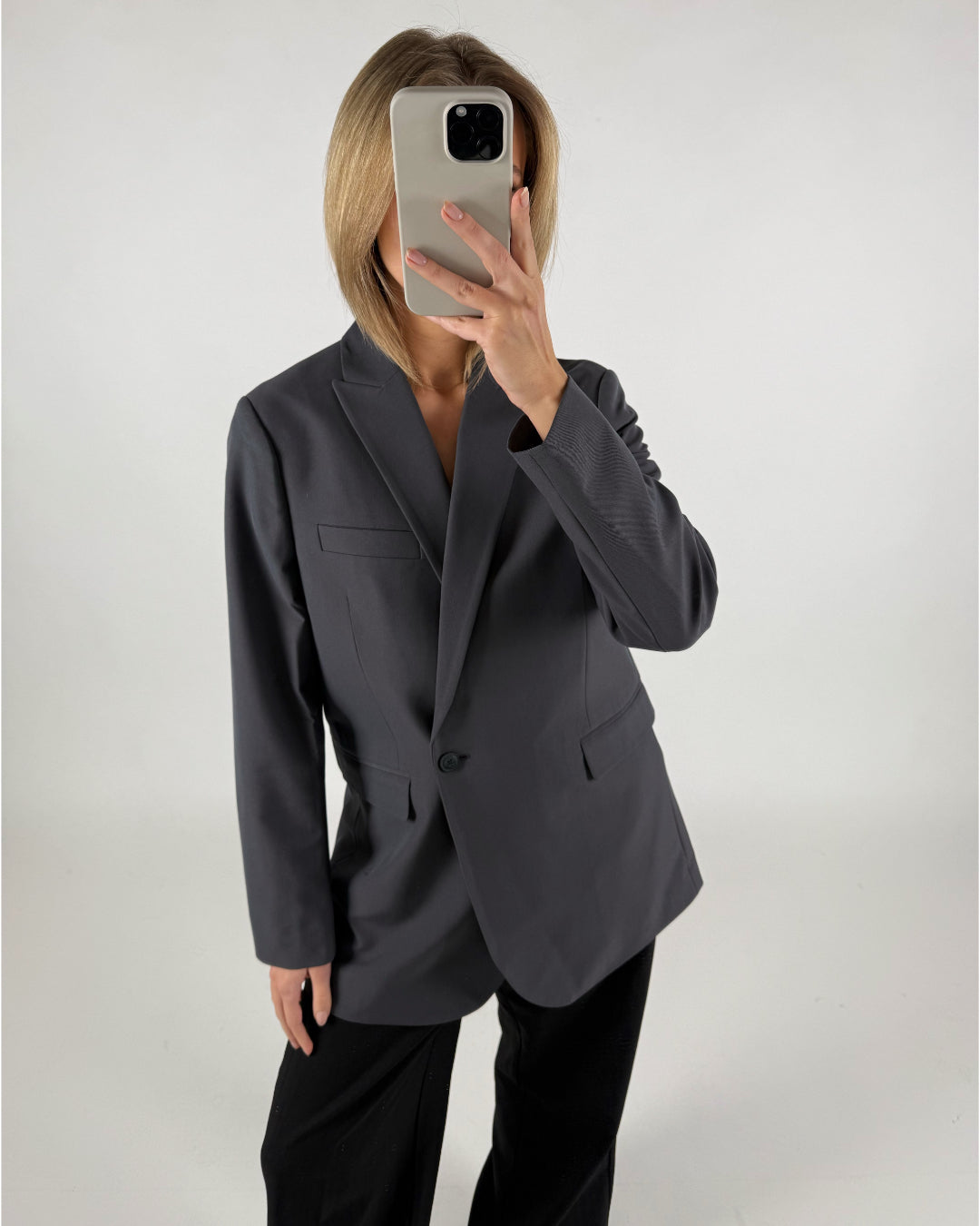 Person iført en mørkegrå blazer holder en smartphone foran ansigtet. Baggrunden er en enkel, lys væg, der fremhæver personens tøj og telefon.