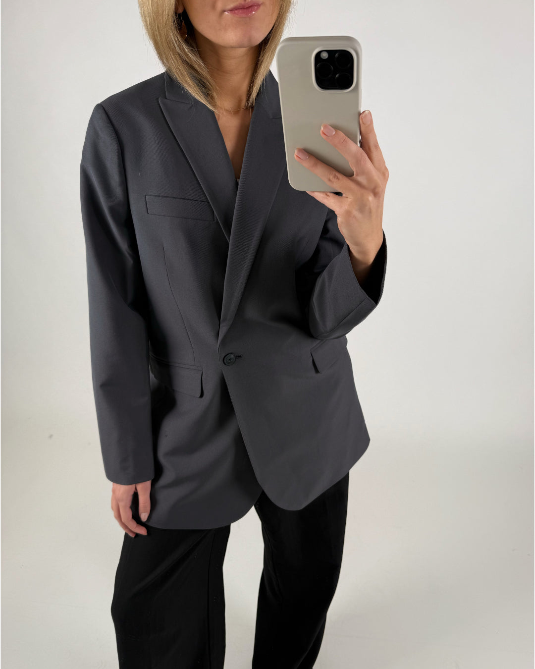 En person iført en mørk blazer tager et spejlbillede med en smartphone. Personen står i et minimalistisk, lyst rum uden synlige dekorationer.