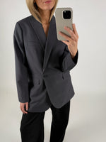 En person iført en mørk blazer tager et spejlbillede med en smartphone. Personen står i et minimalistisk, lyst rum uden synlige dekorationer.