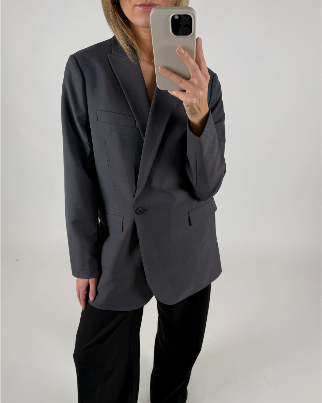 En person iført en mørk blazer tager et selfie med en smartphone. Personen står i et minimalistisk, hvidt rum.