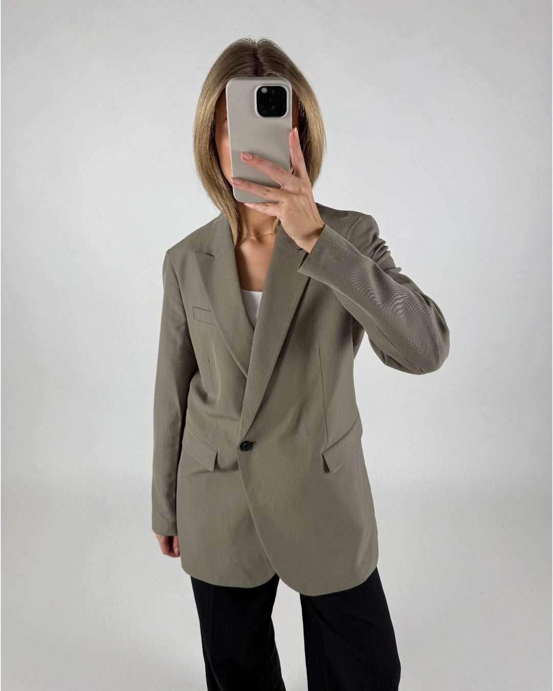 En person iført en grå blazer tager et spejlbillede med en smartphone, stående i et minimalistisk, hvidt rum.