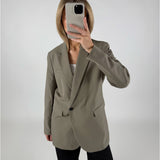 En person iført en grå blazer tager et spejlbillede med en smartphone, stående i et minimalistisk, hvidt rum.