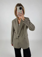 En person iført en grå blazer tager et spejlbillede med en smartphone, stående i et minimalistisk, hvidt rum.