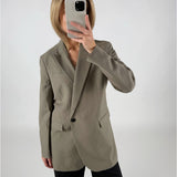 En person iført en grå blazer tager et spejlbillede med en smartphone, der dækker ansigtet, mod en enkel, lys baggrund.