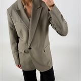 En person iført en beige blazer og sorte bukser tager et spejlbillede med en smartphone i et minimalistisk rum.