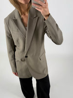 En person iført en beige blazer og sorte bukser tager et spejlbillede med en smartphone i et minimalistisk rum.