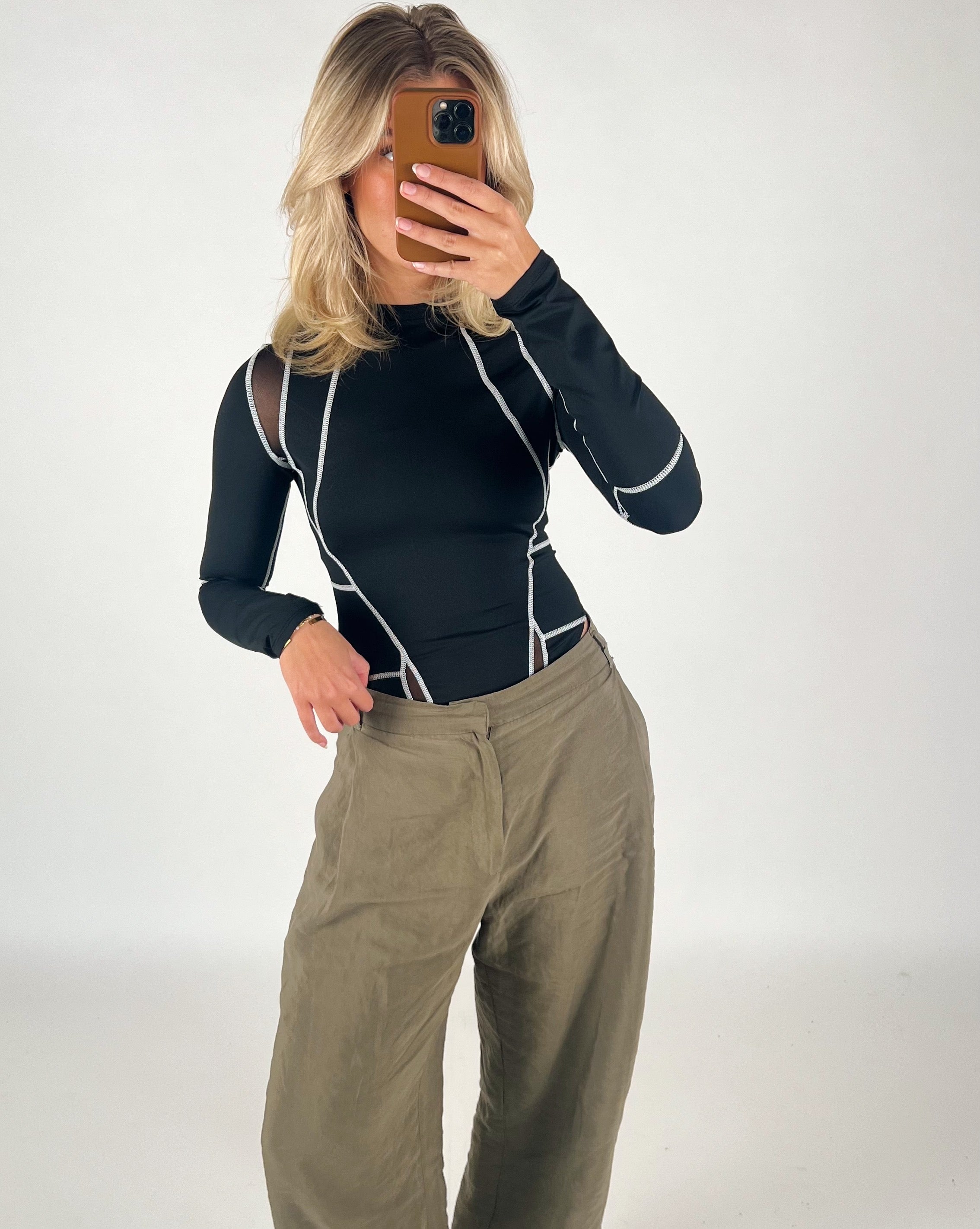 En person tager et spejlbillede iført en sort bluse med hvide linjer og beige bukser, i en simpel indendørs setting. Personen holder en smartphone foran ansigtet.