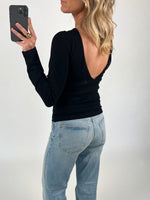 En person iført en sort bluse med åben ryg tager et spejlbillede med en smartphone. De bærer lyseblå jeans i et neutralt oplyst rum.