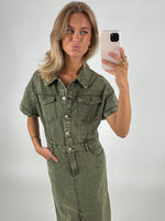 Kvinde iført en olivengrøn denimkjole tager en selfie med sin smartphone i et neutralt, lyst rum. Hun poserer med hovedet let skråt og læberne fremad.