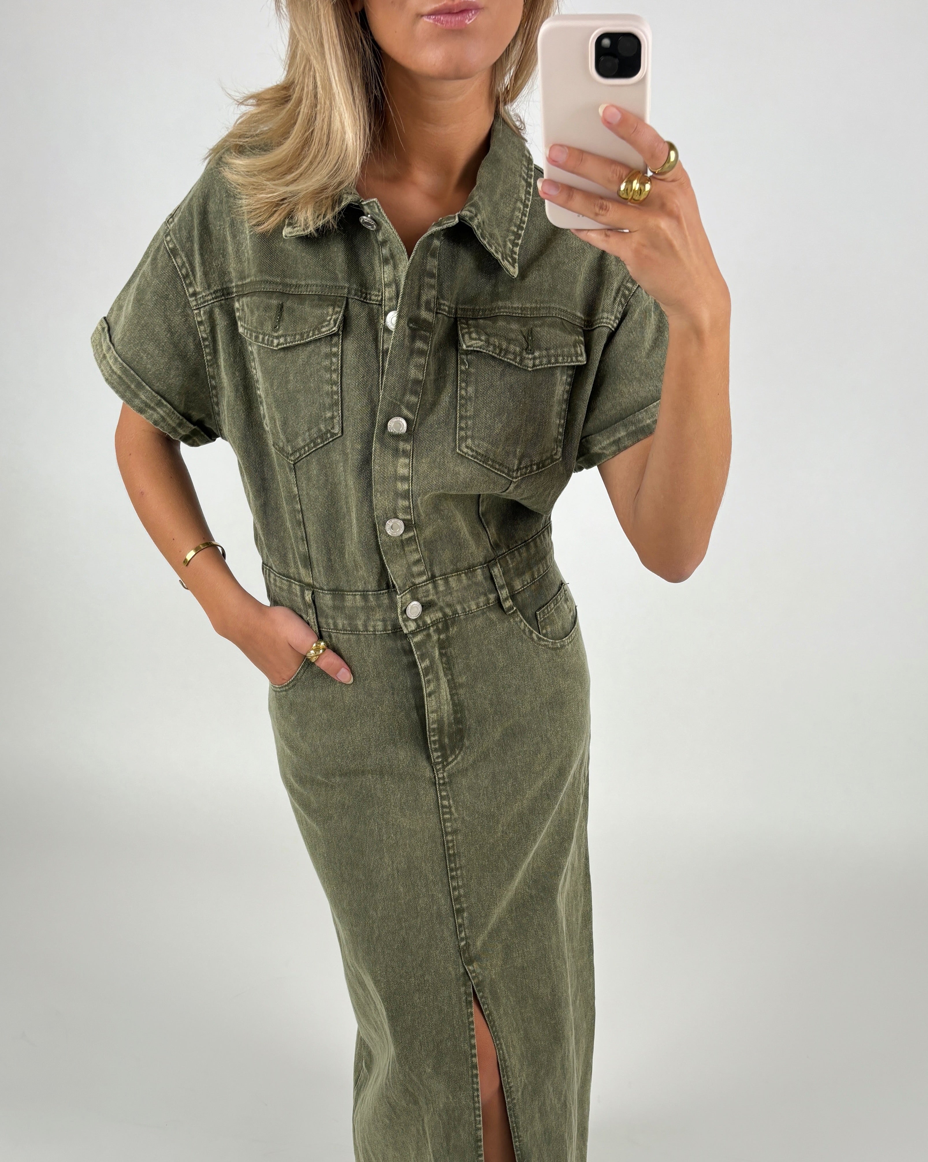 En kvinde iført en grøn denimkjole tager en selfie. Hun står foran en hvid baggrund og har hånden i lommen.