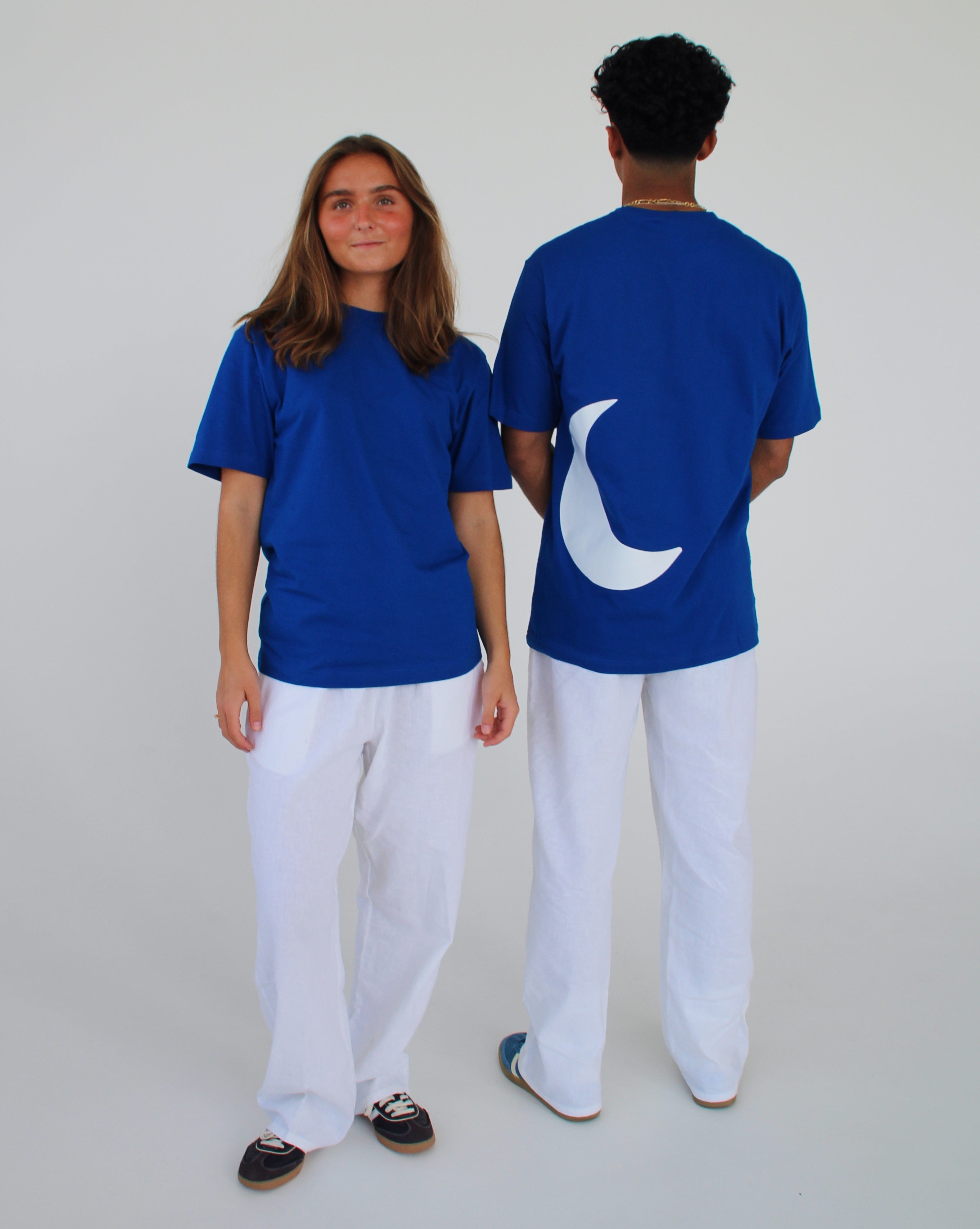 To personer i blå T-shirts og hvide bukser står foran en neutral baggrund; den ene T-shirt har en hvid halvmåne form på ryggen.