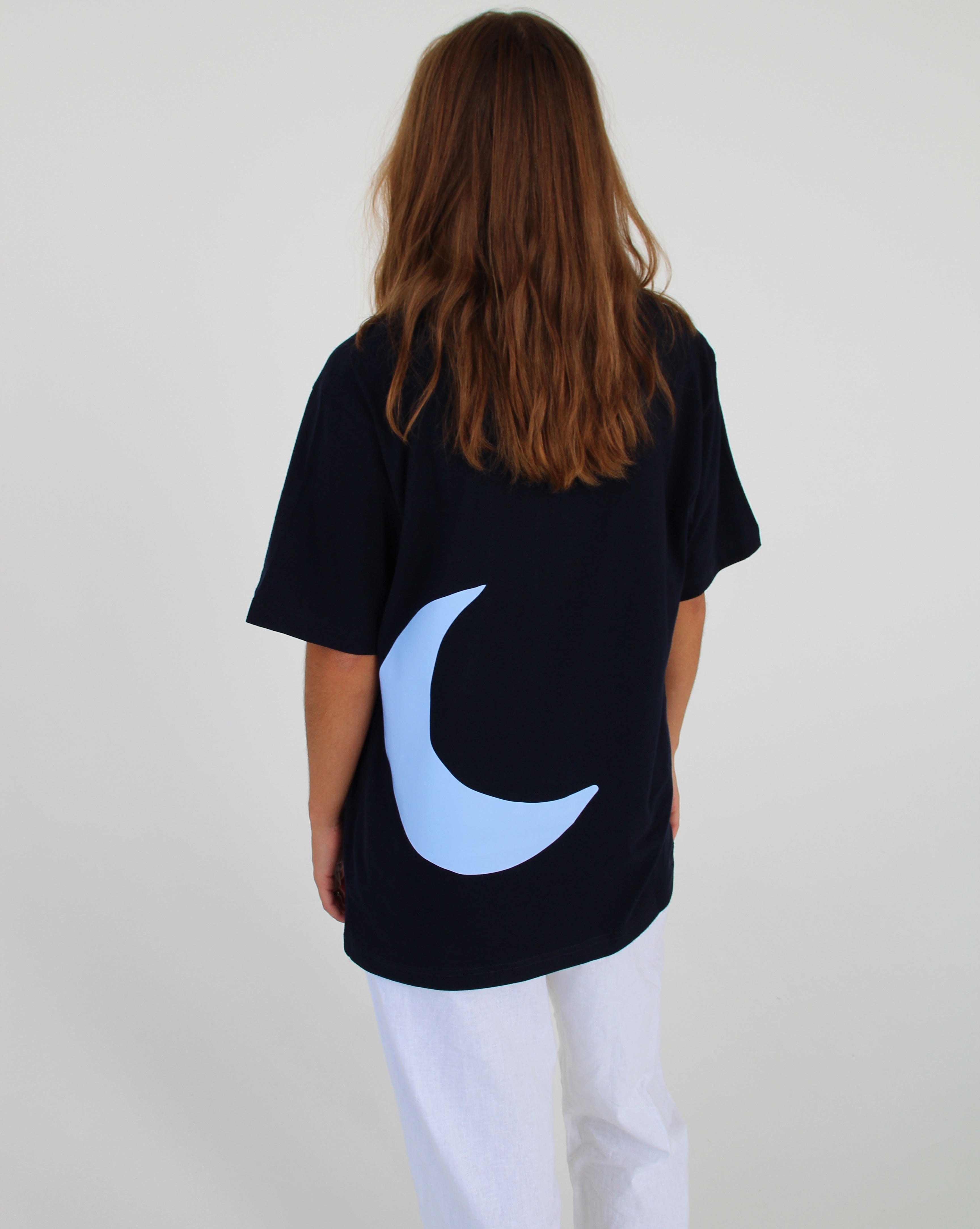 En person iført en sort T-shirt med en blå måne på ryggen står med ryggen til og har langt, brunt hår. De har hvide bukser på i et neutralt rum.