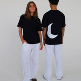 To personer iført sorte T-shirts og hvide bukser står i et hvidt rum; den ene er vendt mod kameraet, mens den anden viser en hvid halvmåne på ryggen.