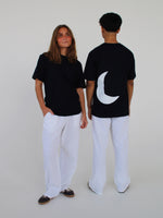 To personer iført sorte T-shirts og hvide bukser står i et hvidt rum; den ene er vendt mod kameraet, mens den anden viser en hvid halvmåne på ryggen.