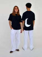 To personer i sorte t-shirts og hvide bukser står sammen; den ene vender mod kameraet, den anden med ryggen til. T-shirten har en stor hvid halvmåne bagpå.