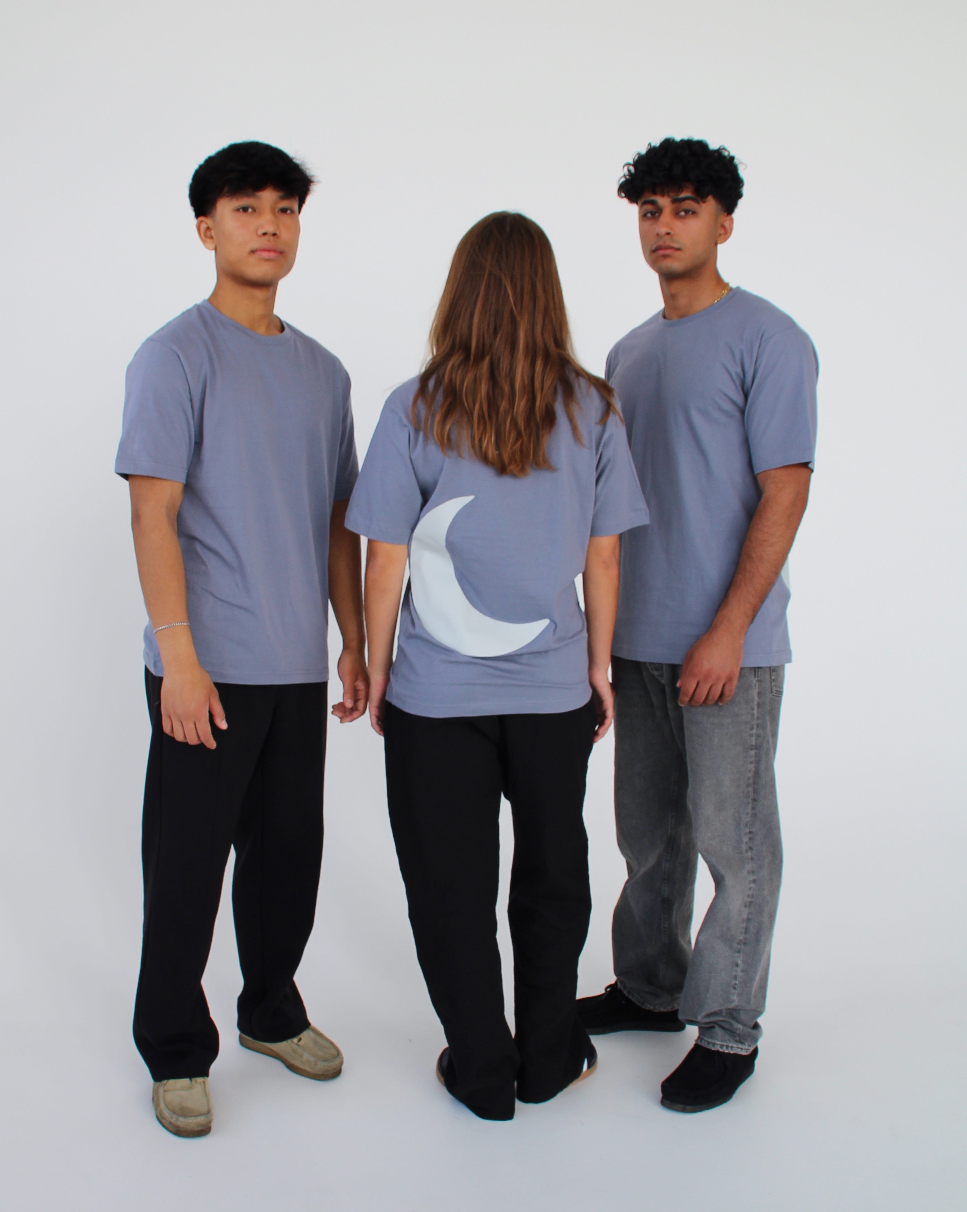 Tre personer står i en lys baggrund. To mænd i grå T-shirts vender mod kameraet, og en kvinde i lignende T-shirt med en hvid måne på ryggen står med ryggen til.