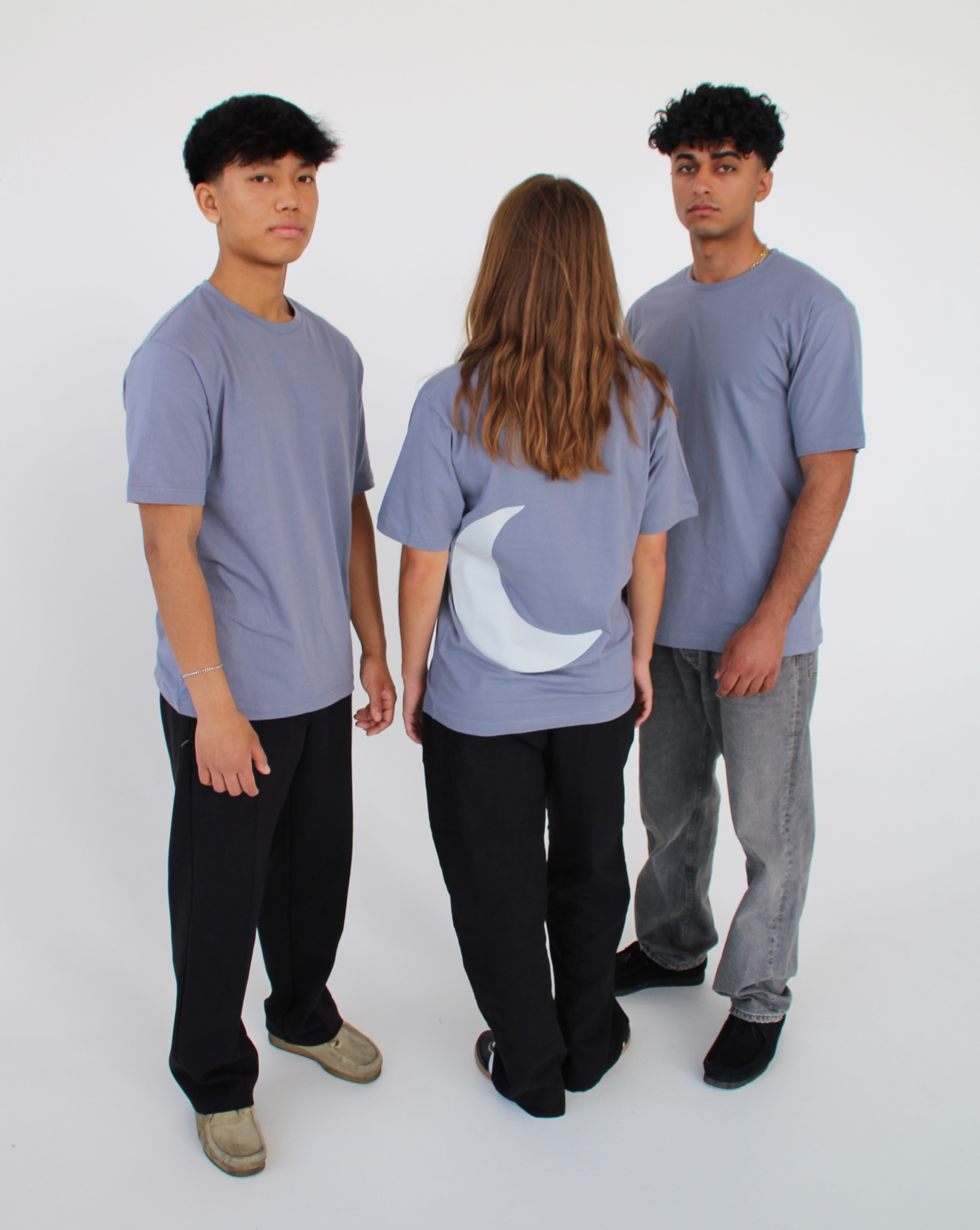 Tre personer står sammen i en cirkel, iført grå t-shirts. En person vender ryggen til, med en måneform på trøjen. De er i et minimalistisk, hvidt rum.
