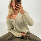 En person sidder på gulvet med krydsede ben, iført en beige sweater og khakifarvede bukser. Personen holder en smartphone op foran ansigtet i et lyst rum.