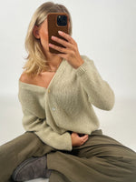 En person sidder på gulvet med krydsede ben, iført en beige sweater og khakifarvede bukser. Personen holder en smartphone op foran ansigtet i et lyst rum.