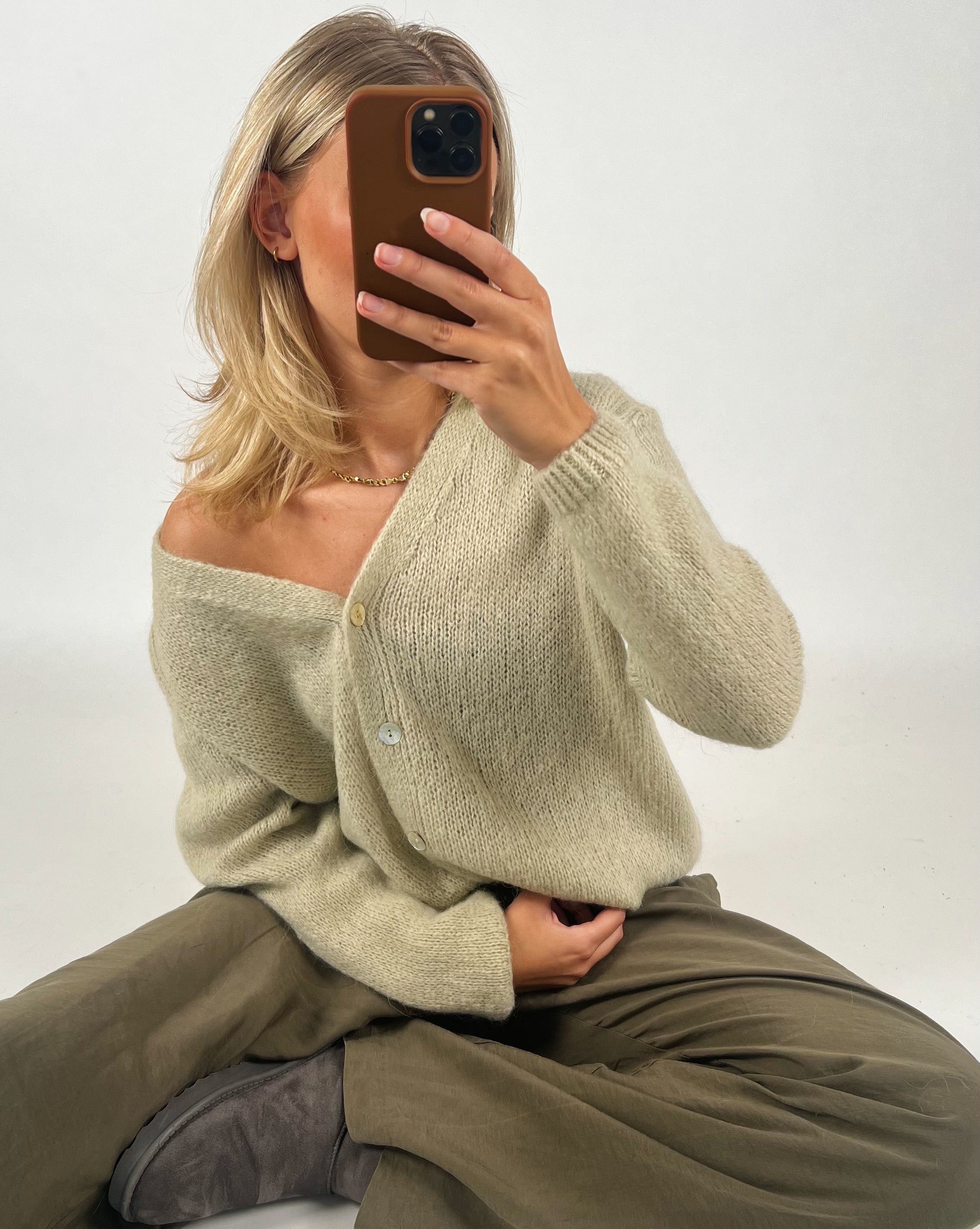 En person sidder på gulvet med krydsede ben, iført en beige sweater og khakifarvede bukser. Personen holder en smartphone op foran ansigtet i et lyst rum.