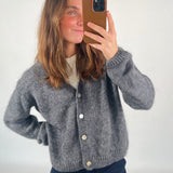 En person iført en grå cardigan tager en selfie med en smartphone i et lysegråt rum. Cardiganen har hvide knapper, og personen har en hånd i siden.