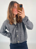 En person iført en grå cardigan tager en selfie med en smartphone i et lysegråt rum. Cardiganen har hvide knapper, og personen har en hånd i siden.