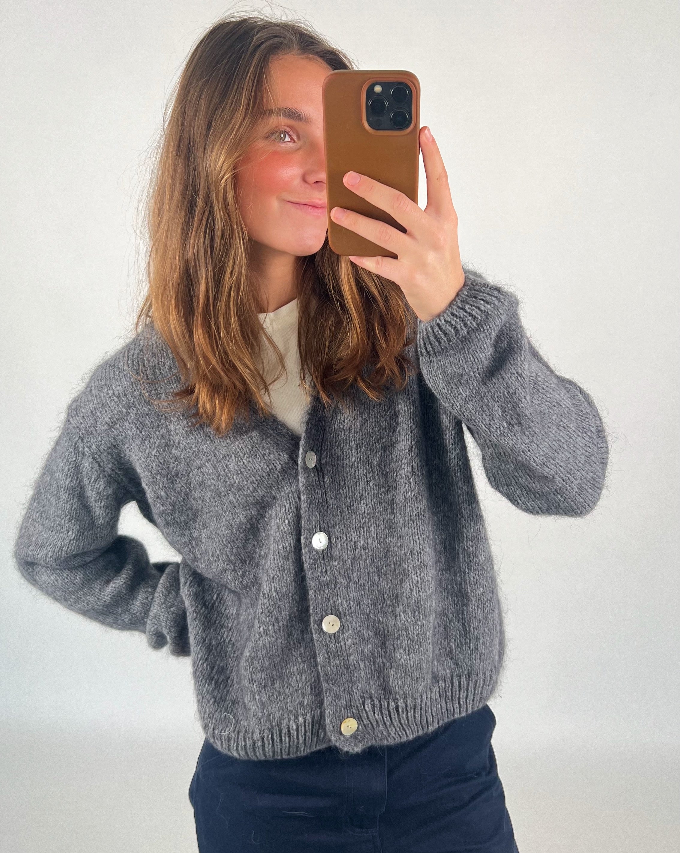 En person iført en grå cardigan tager en selfie med en smartphone i et lysegråt rum. Cardiganen har hvide knapper, og personen har en hånd i siden.