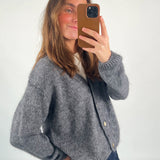 En person tager et spejlbillede med en smartphone. Vedkommende bærer en grå cardigan over en hvid top, i et neutralt oplyst rum.