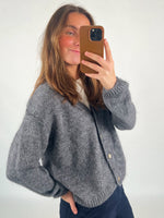En person tager et spejlbillede med en smartphone. Vedkommende bærer en grå cardigan over en hvid top, i et neutralt oplyst rum.