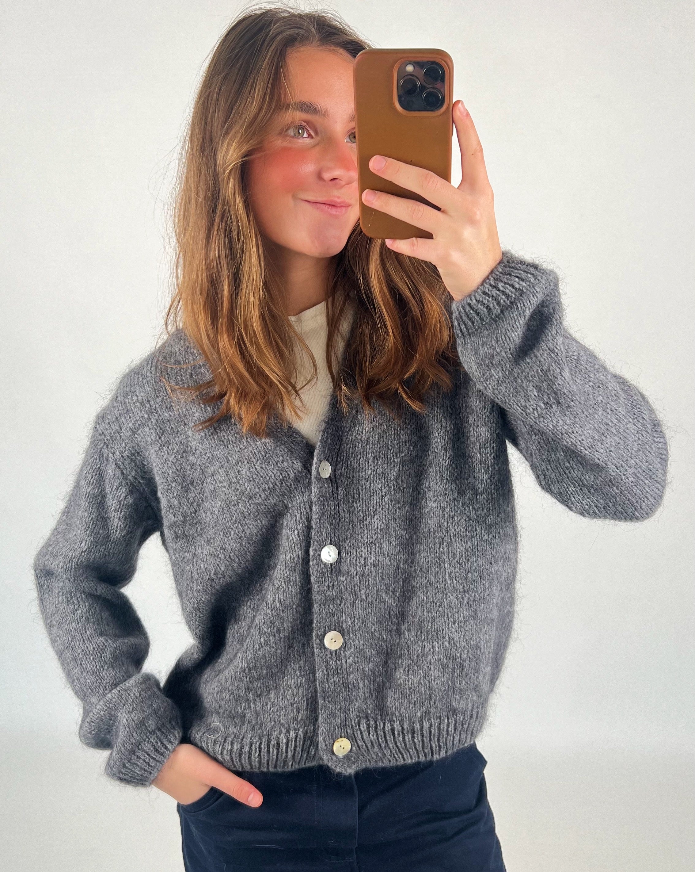 En person iført en grå cardigan tager en selfie med en brun mobiltelefon i et lyst rum. Personen har hånden i bukselommen.