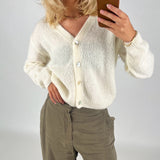 En person iført en hvid cardigan tager en selfie med en brun mobiltelefon i et neutralt rum. Personen bærer også grå bukser og en guldhalskæde.