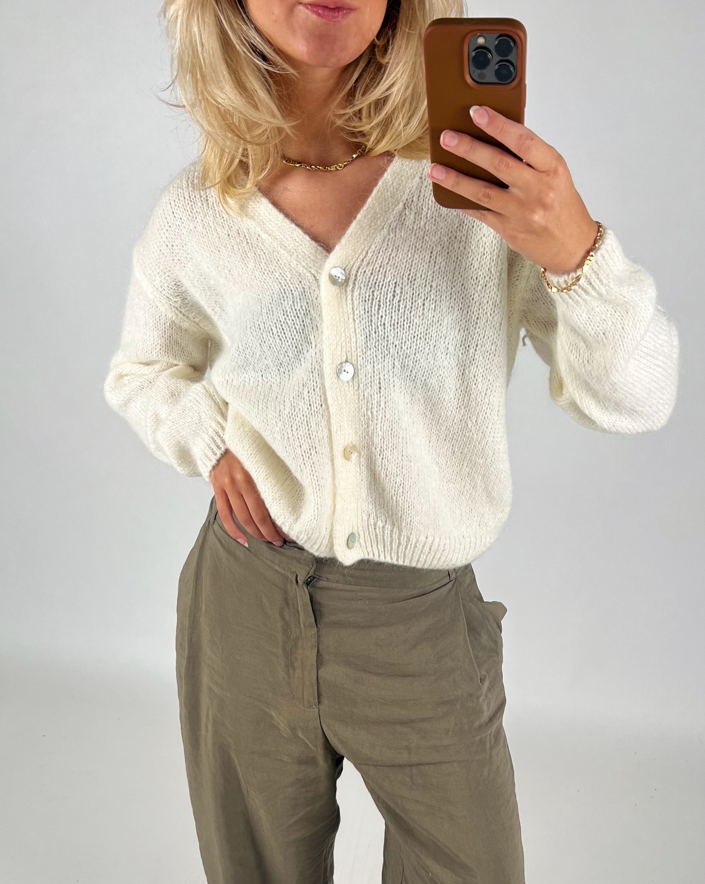 En person iført en hvid cardigan tager en selfie med en brun mobiltelefon i et neutralt rum. Personen bærer også grå bukser og en guldhalskæde.