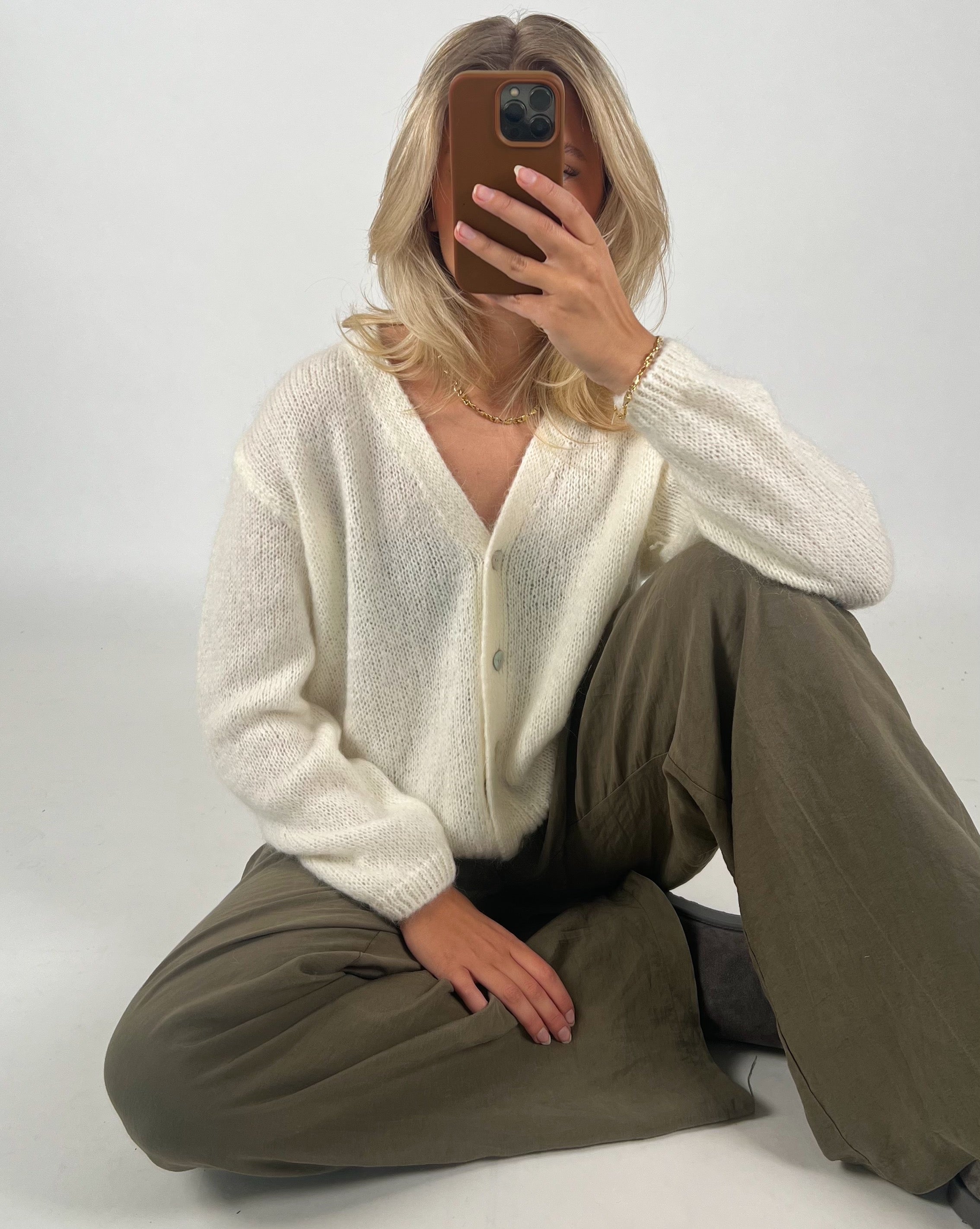 En person iført en hvid sweater og olivenfarvede bukser sidder på gulvet og dækker ansigtet med en mobiltelefon. Baggrunden er neutral og lysegrå.