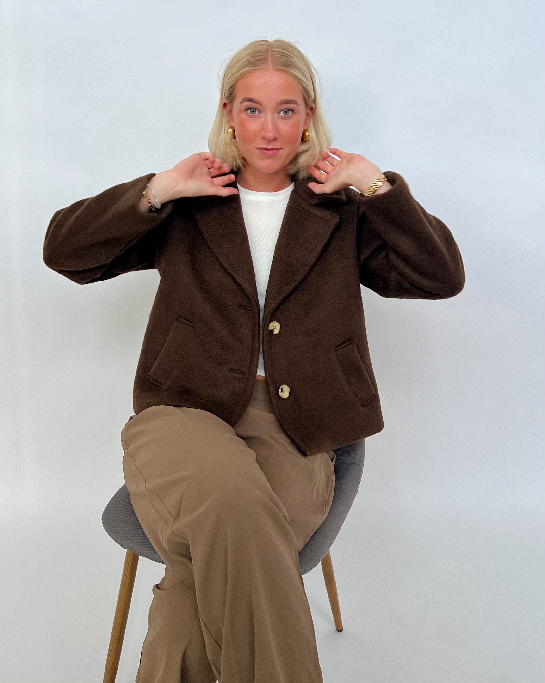 En person sidder på en stol og justerer sin brune jakke. De har beige bukser på og er i et neutralt rum.