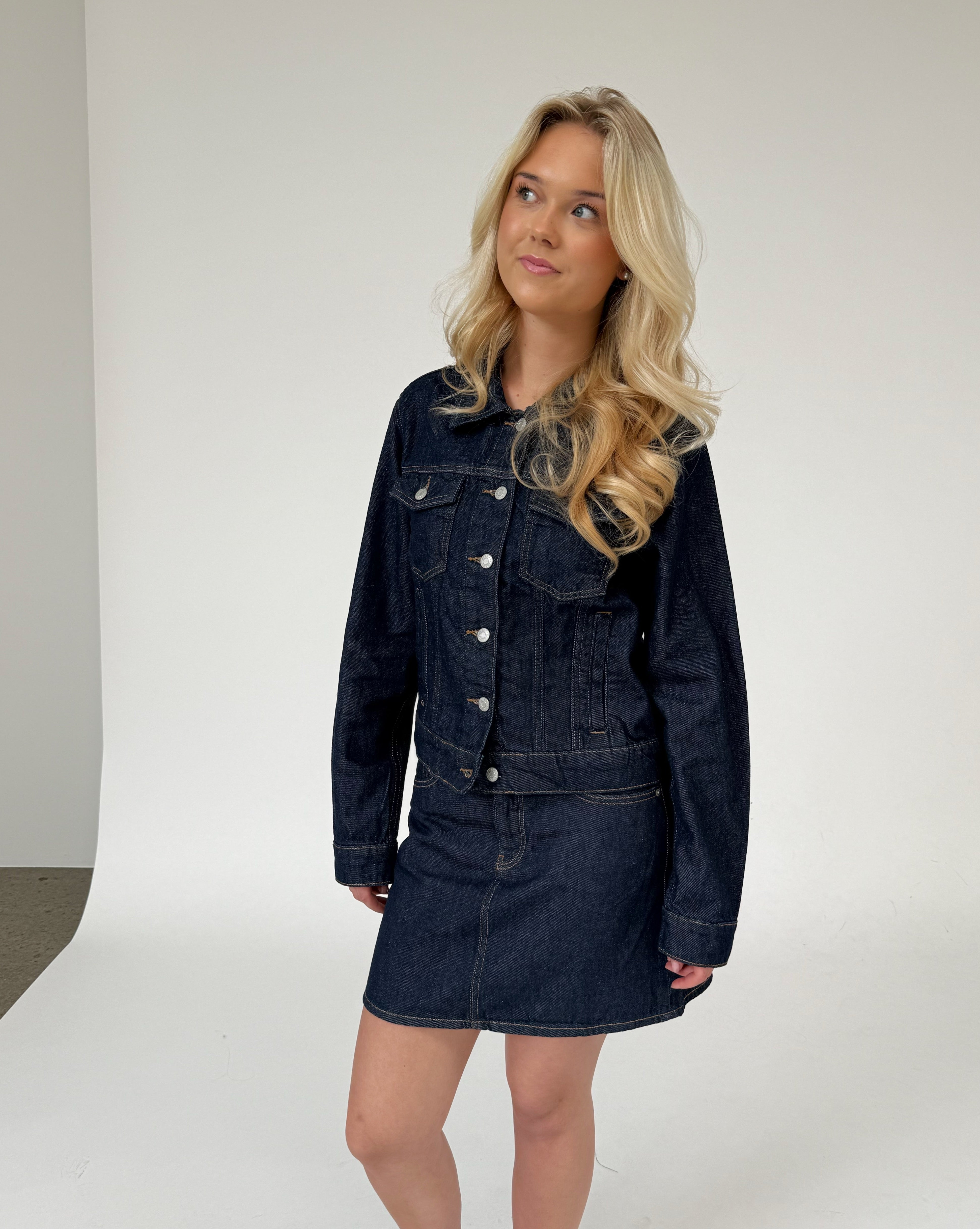 En person med lyst hår iført en mørk denimjakke og nederdel står og ser opad i et fotostudie med en hvid baggrund.