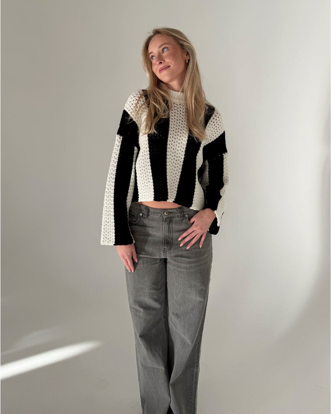 Person iført sort og hvid stribet sweater og grå bukser står afslappet i et minimalistisk rum med en lys væg i baggrunden.