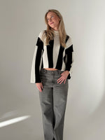 Person iført sort og hvid stribet sweater og grå bukser står afslappet i et minimalistisk rum med en lys væg i baggrunden.