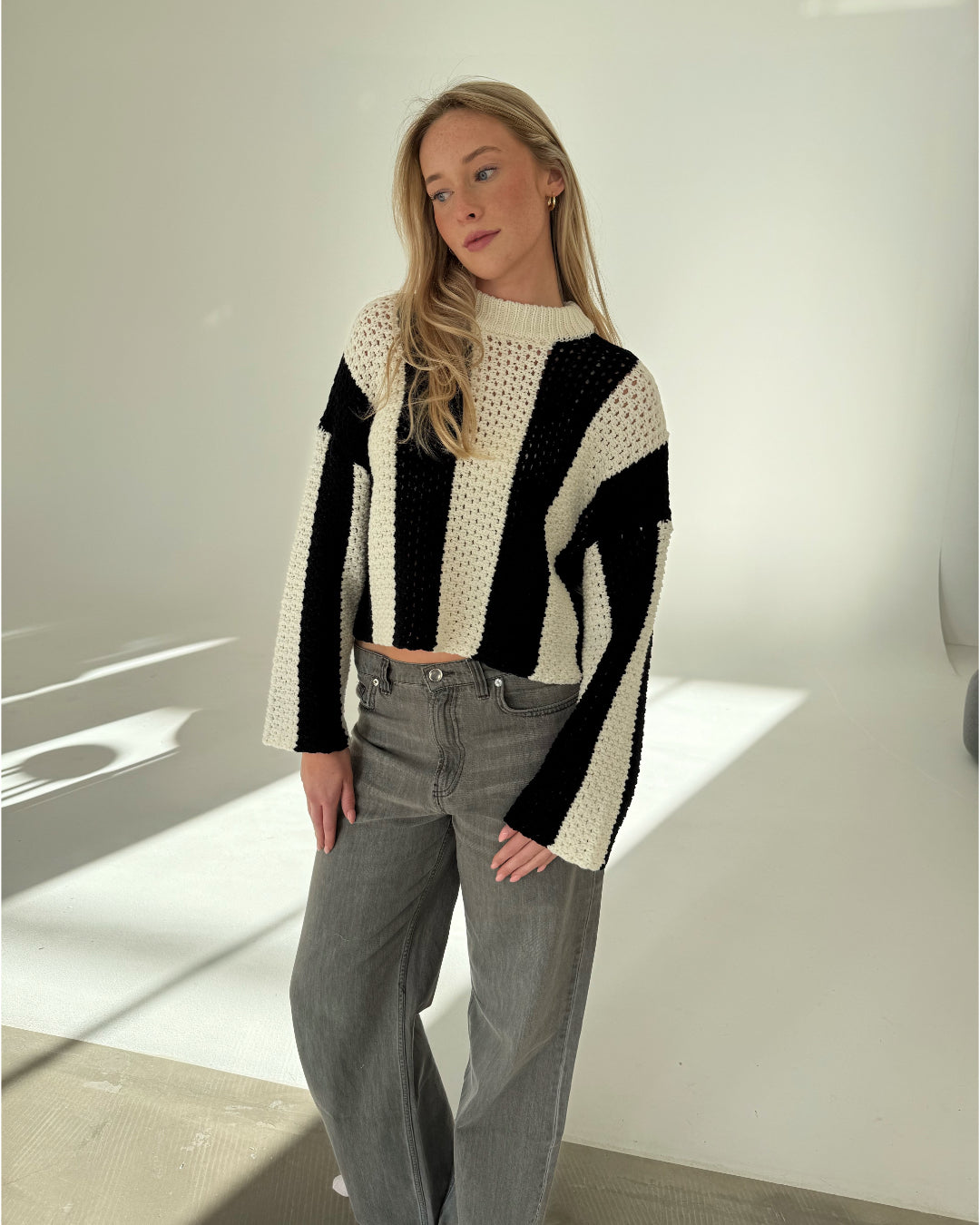 En person iført en stribet, sort-hvid sweater står afslappet i et lyst rum med gråt gulv, med hænderne på hofterne og kigger til siden.