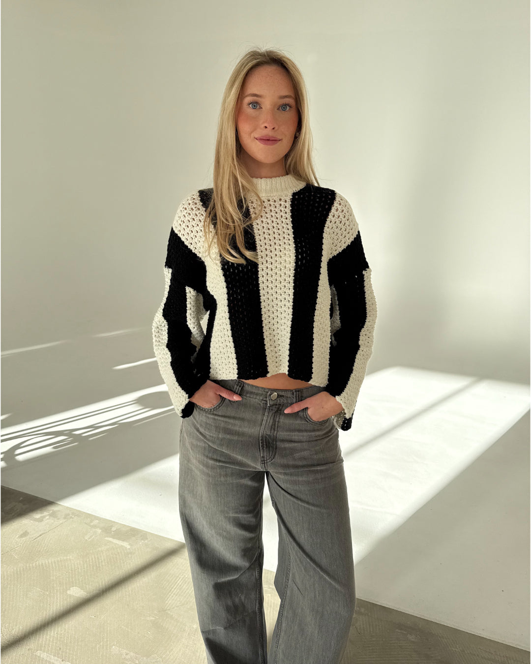 Kvinde iført stribet sweater står med hænderne i lommerne i et lyst rum med lysegråt gulv. Hun kigger mod kameraet, og lyset skaber skygger på gulvet.