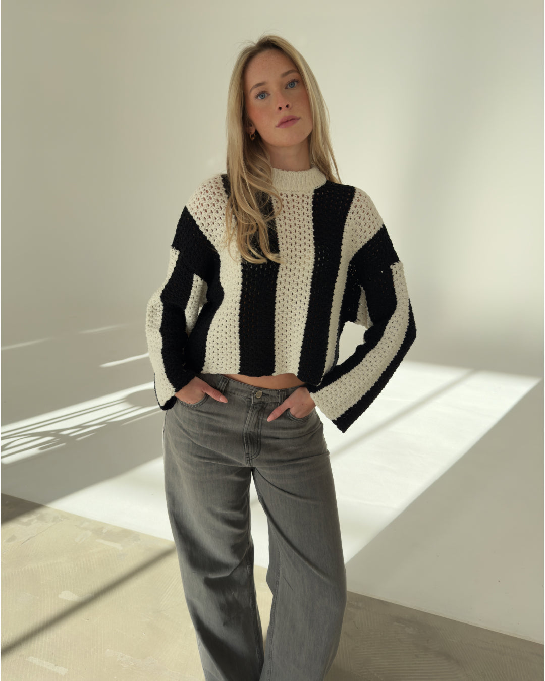 En person står stille, iført en sort-hvid stribet striktrøje og grå jeans, i et rum med lys, der skaber skygger på væggen.